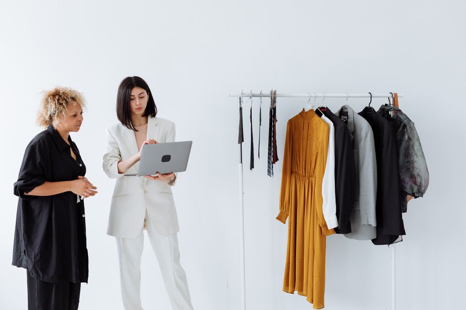Two fashion designers discussing clothing options with a laptop in a boutique setting.